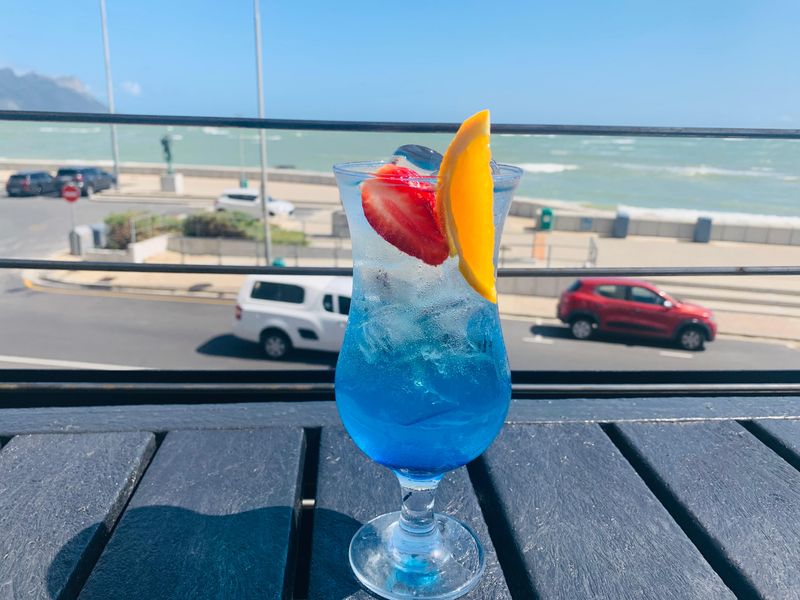 A blue cocktail with fruit garnish beside a window overlooking Strand beach